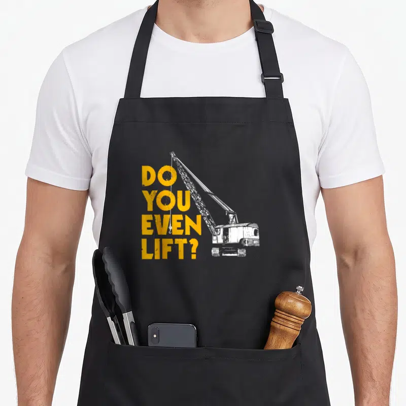 Geschenke construction apron - Yellow stacked text DO YOU EVEN LIFT next to a white crane with hanging hook — shown on a black cotton canvas apron against a white background