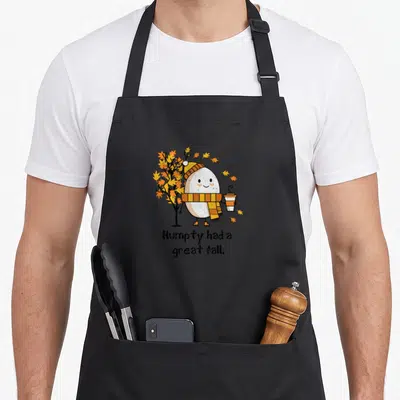 Distressed cute fall aprons - Egg character in scarf holding coffee next to autumn tree with Humpty had a great fall — shown on a black cotton canvas apron against a white background