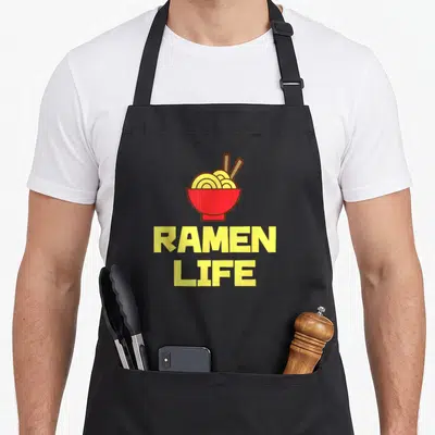 Ramen tasty aprons - Red bowl with yellow noodles and RAMEN LIFE text in cartoon style — shown on a black cotton canvas apron against a white background