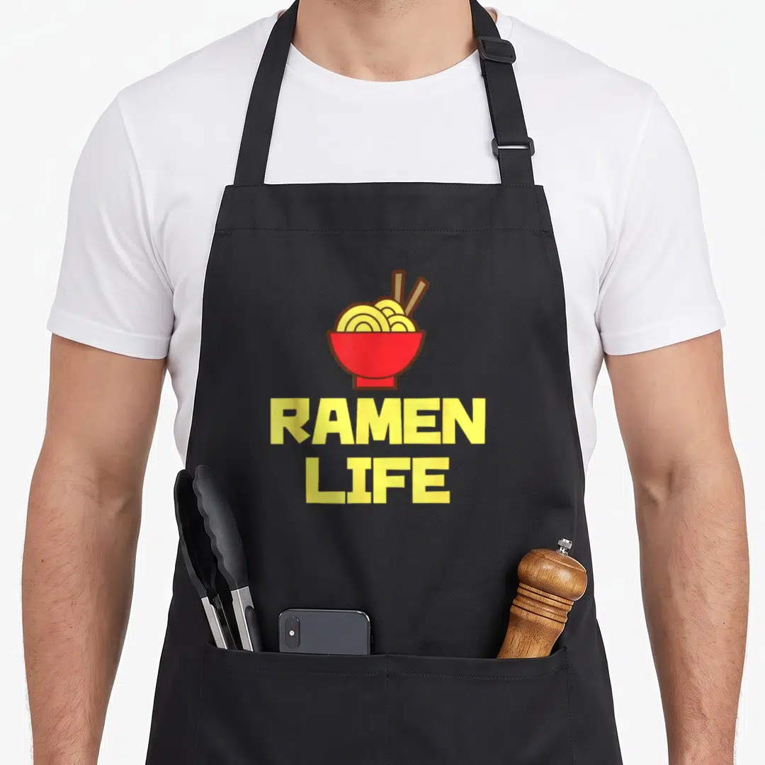 Ramen tasty aprons - Red bowl with yellow noodles and RAMEN LIFE text in cartoon style — shown on a black cotton canvas apron against a white background