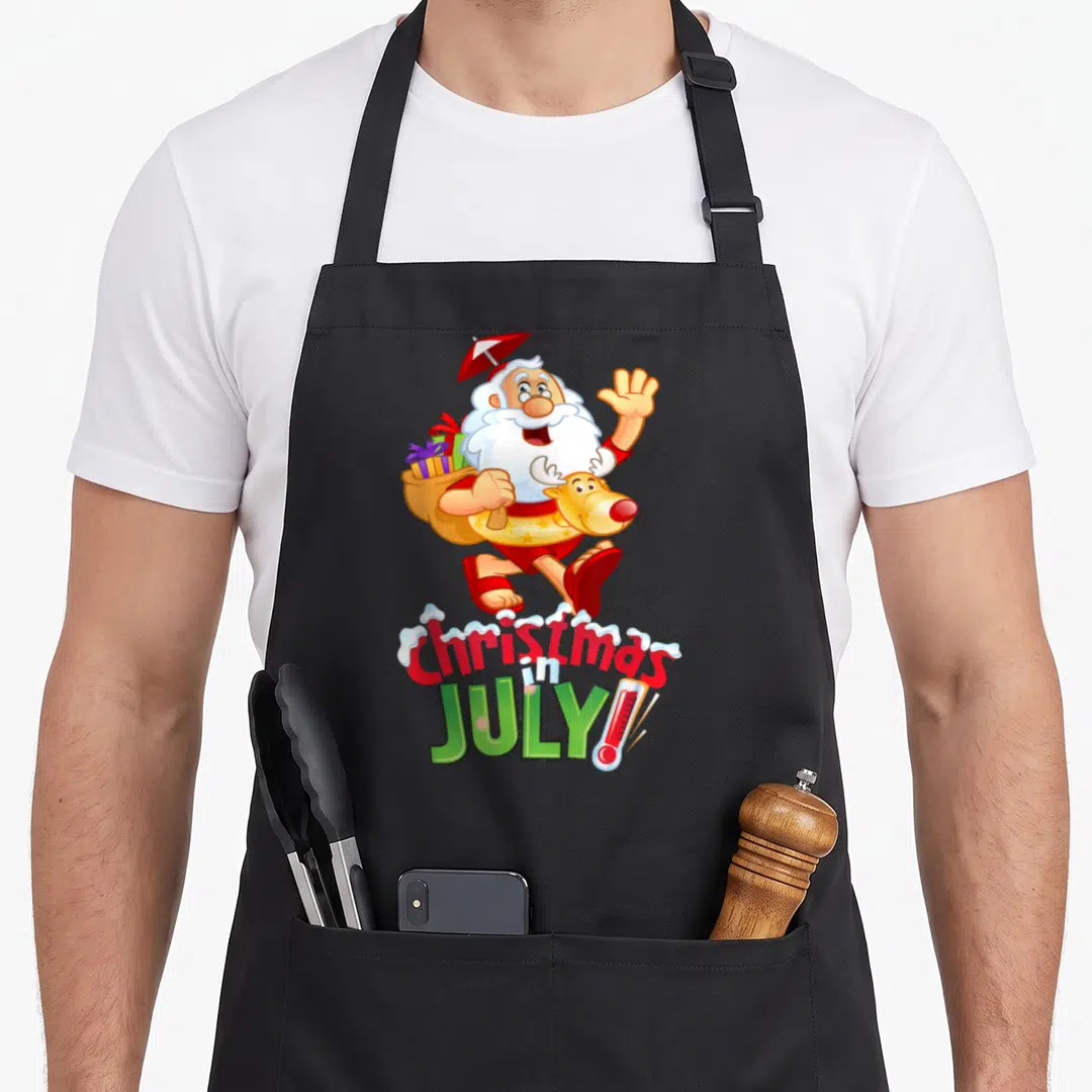 July reindeer christmas apron - Santa in swimwear holding umbrella and sack with Christmas in July text and thermometer — shown on a black cotton canvas apron against a white background