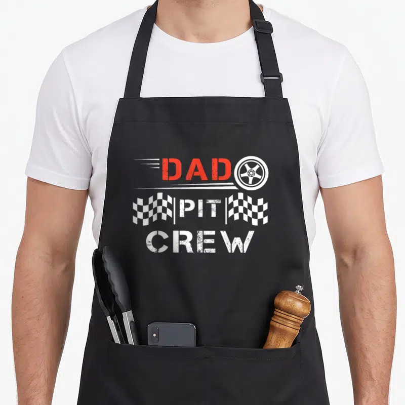 Kart daddy apron - Stacked DAD PIT CREW text with wheel and checkered flags in red and white — shown on a black cotton canvas apron against a white background