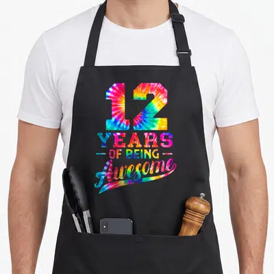 12th tie apron - Tie-dye text reading 12 Years of Being Awesome with pink blue yellow green colors and swoosh — shown on a black cotton canvas apron against a white background