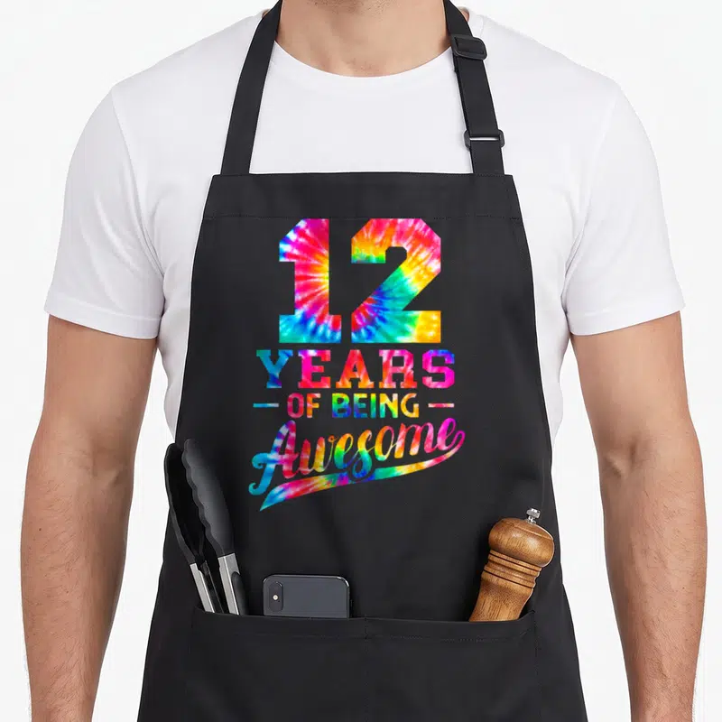 12th tie apron - Tie-dye text reading 12 Years of Being Awesome with pink blue yellow green colors and swoosh — shown on a black cotton canvas apron against a white background