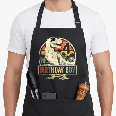 4th birthday t rex apron - Cartoon T-Rex in sunglasses with BIRTHDAY BOY text and retro sun design — shown on a black cotton canvas apron against a white background