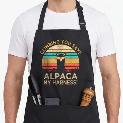 Climbing alpaca apron - Retro striped sun with grey alpaca wearing orange sunglasses and climbing pun text — shown on a black cotton canvas apron against a white background