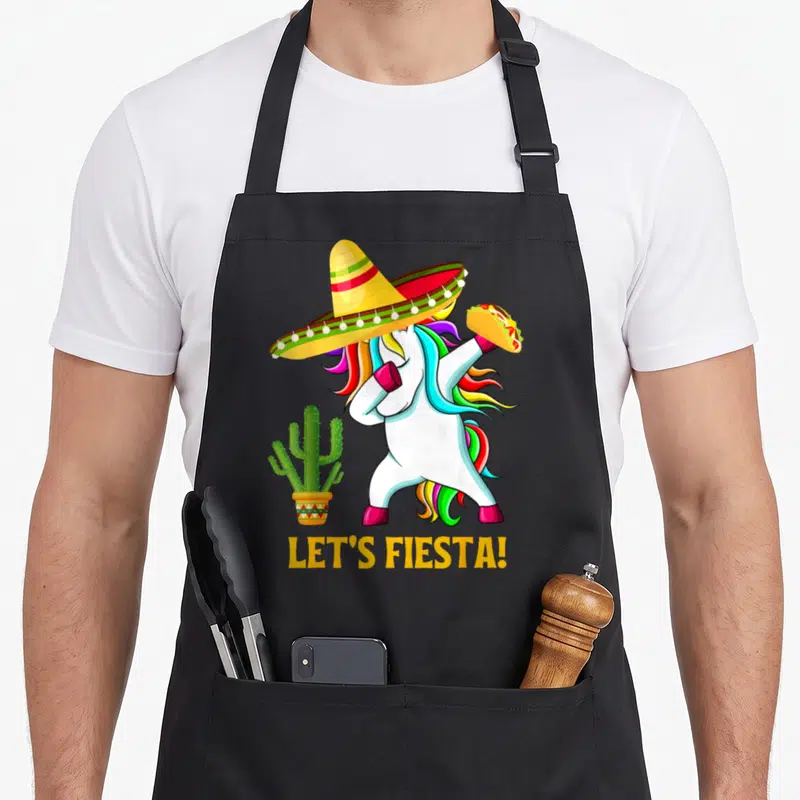 Cinco unicorn apron - Cartoon white unicorn dabbing in sombrero holding taco next to cactus with LET'S FIESTA text — shown on a black cotton canvas apron against a white background