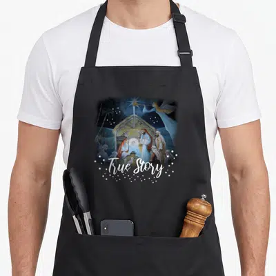 Story christian womens christmas apron - Nativity scene with angel and True Story script surrounded by white stars — shown on a black cotton canvas apron against a white background