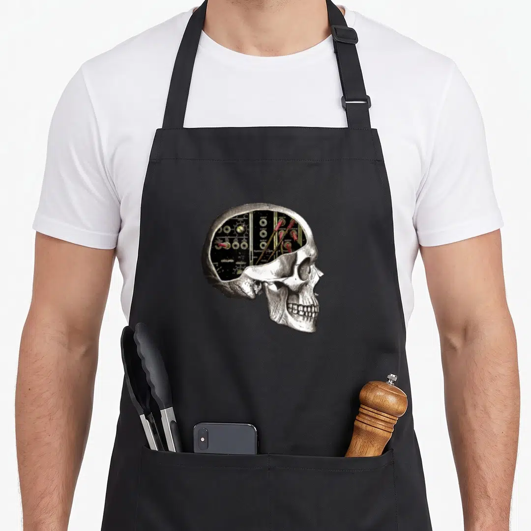 Analog skull apron - Realistic skull with analog synth panel inside cranium showing output jacks and oscillator labels — shown on a black cotton canvas apron against a white background
