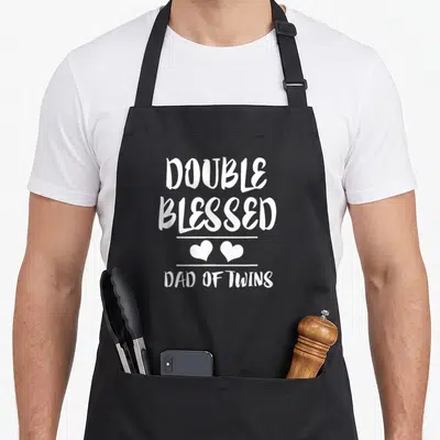 Father double apron - White stacked text Double Blessed Dad of Twins with two hearts between horizontal lines — shown on a black cotton canvas apron against a white background