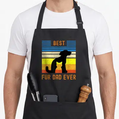 Fur funny cat aprons - Best Fur Dad Ever text with black dog silhouette holding orange cat on colorful striped — shown on a black cotton canvas apron against a white background