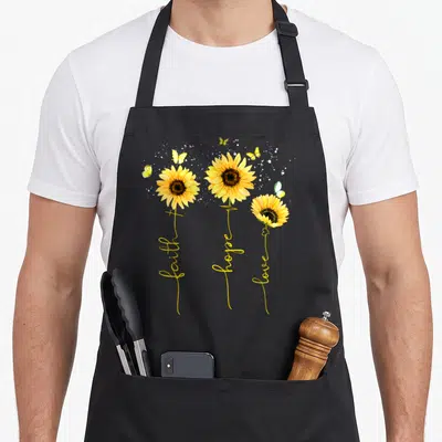 Hope faith aprons - Three sunflowers with stems spelling faith hope love in yellow cursive with butterflies — shown on a black cotton canvas apron against a white background