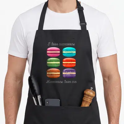 Macaron cupcake apron - Six colorful cartoon macarons in pink blue green red orange purple between black script text — shown on a black cotton canvas apron against a white background