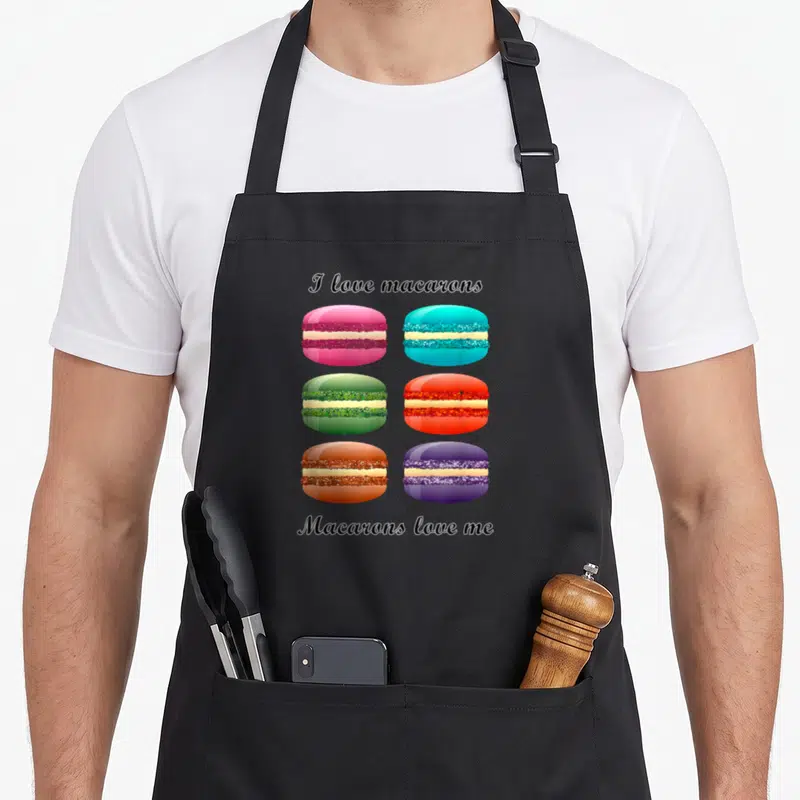 Macaron cupcake apron - Six colorful cartoon macarons in pink blue green red orange purple between black script text — shown on a black cotton canvas apron against a white background