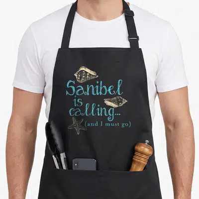 Sanibel florida apron - Blue script text with beige seashells and starfish line art on dark background — shown on a black cotton canvas apron against a white background