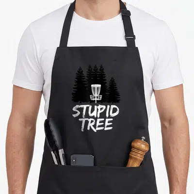Stupid golf apron - White disc golf basket in front of black pine trees with STUPID TREE text — shown on a black cotton canvas apron against a white background