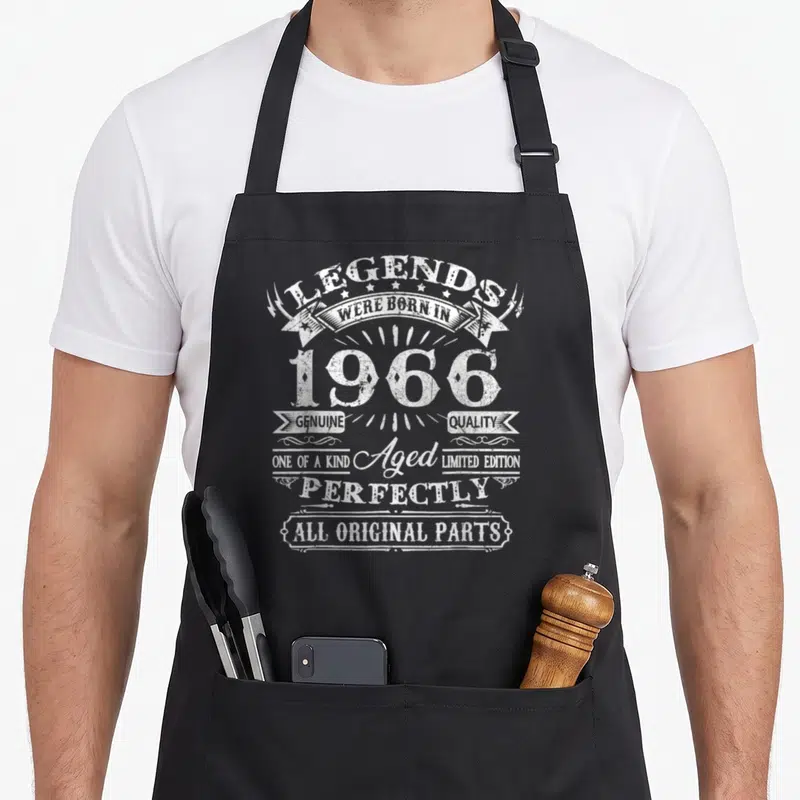 Funny legend 60th birthday apron - White distressed text reading Legends Were Born In 1966 with stars and ribbons — shown on a black cotton canvas apron against a white background