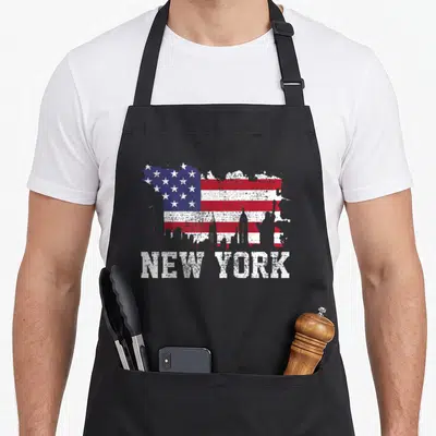 Flag new york apron - Distressed USA flag paint splatter with white skyline silhouette and NEW YORK text below — shown on a black cotton canvas apron against a white background