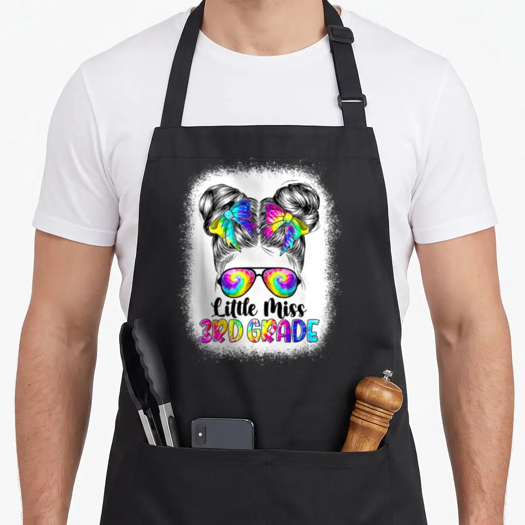 Miss 3rd tie-dye apron - Cartoon girl head with two buns, tie-dye sunglasses and Little Miss 3rd Grade text in black — shown on a black cotton canvas apron against a white background