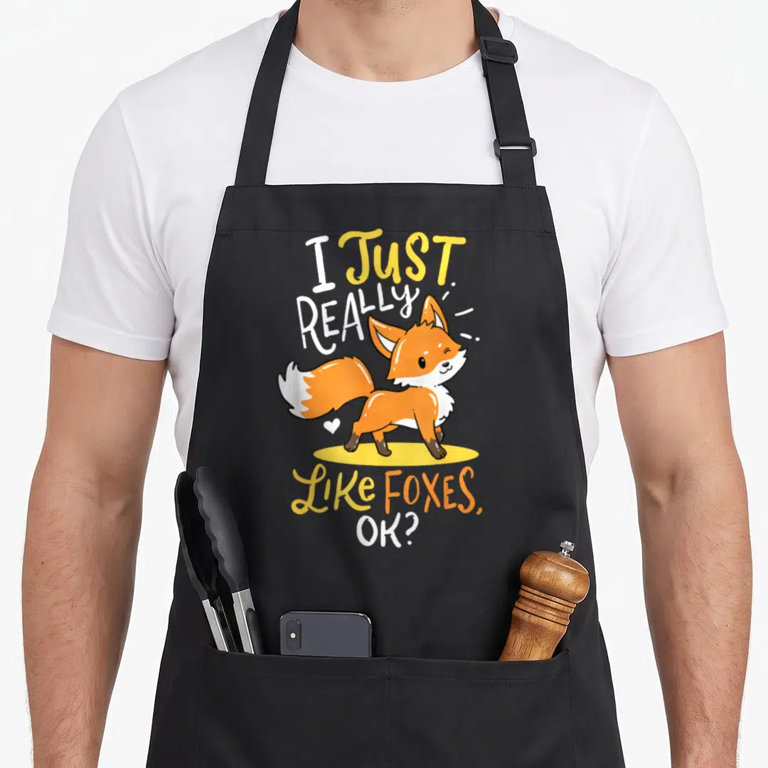 Baby smart apron - Cartoon orange fox on yellow oval with I Just Really Like Foxes Ok text and small white heart — shown on a black cotton canvas apron against a white background