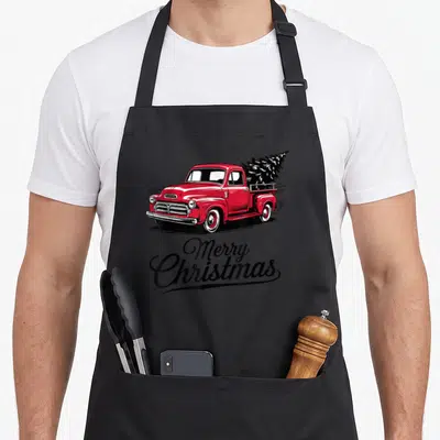 Pickup truck christmas tree apron - Red vintage truck carrying black Christmas tree above Merry Christmas script — shown on a black cotton canvas apron against a white background