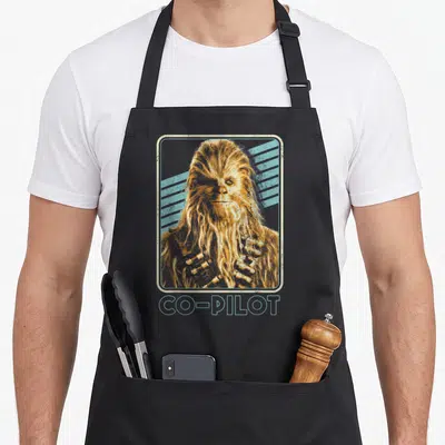 Chewie pilot chef and co aprons - Distressed Chewbacca portrait in rounded frame with diagonal stripes and CO-PILOT — shown on a black cotton canvas apron against a white background