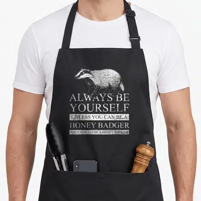 Honey badger apron - Distressed white honey badger illustration above stacked text always be yourself unless you can be — shown on a black cotton canvas apron against a white background