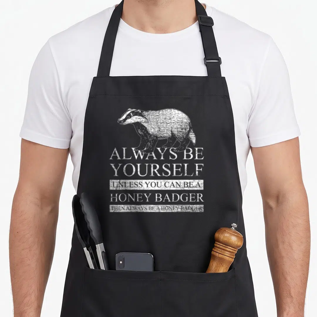 Honey badger apron - Distressed white honey badger illustration above stacked text always be yourself unless you can be — shown on a black cotton canvas apron against a white background