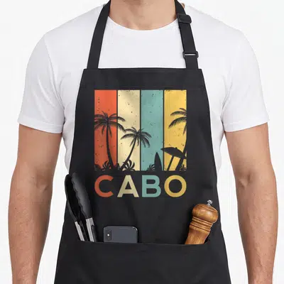 Cabo mexico apron - Retro beach scene with palm trees, surfboard, and CABO text over four colored stripes — shown on a black cotton canvas apron against a white background
