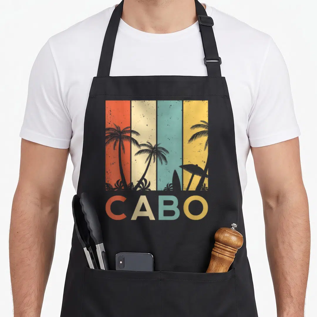 Cabo mexico apron - Retro beach scene with palm trees, surfboard, and CABO text over four colored stripes — shown on a black cotton canvas apron against a white background