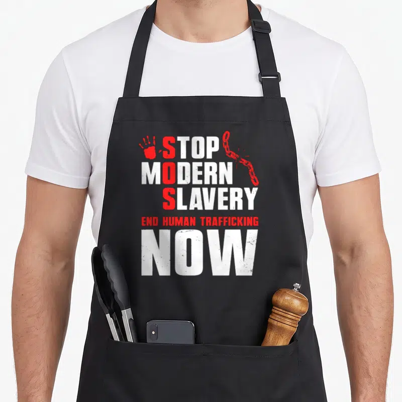Slavery modern apron - Stacked white and red text reading STOP MODERN SLAVERY with red handprint and broken chain — shown on a black cotton canvas apron against a white background
