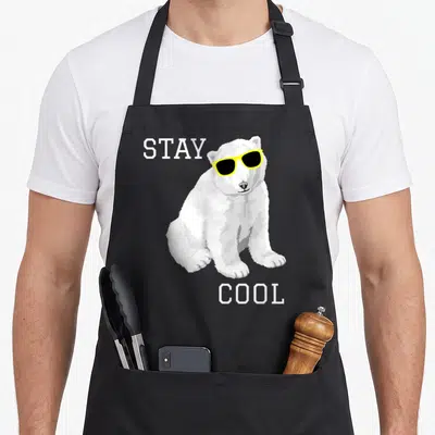 Stay bear apron - Cartoon polar bear with yellow sunglasses and STAY COOL text — shown on a black cotton canvas apron against a white background