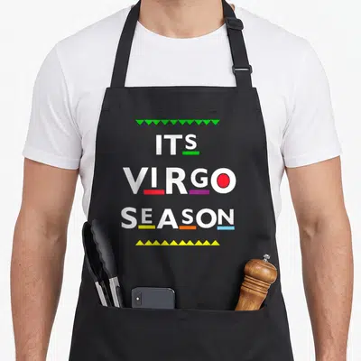 Virgo season birthday apron - ITS VIRGO SEASON text with green top triangles, yellow bottom triangles, and colorful — shown on a black cotton canvas apron against a white background