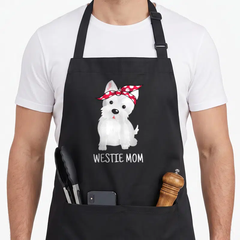Terrier westie apron - Cartoon white dog wearing red polka dot bandana with text WESTIE MOM below — shown on a black cotton canvas apron against a white background