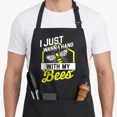 Hang bee apron - I Just Wanna Hang With My Bees text with cartoon bee inside yellow hexagon — shown on a black cotton canvas apron against a white background