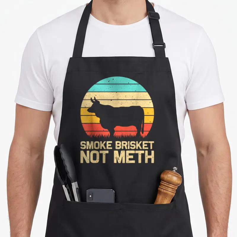 Smoke brisket apron - Grey cow silhouette over retro striped sun with text SMOKE BRISKET NOT METH — shown on a black cotton canvas apron against a white background