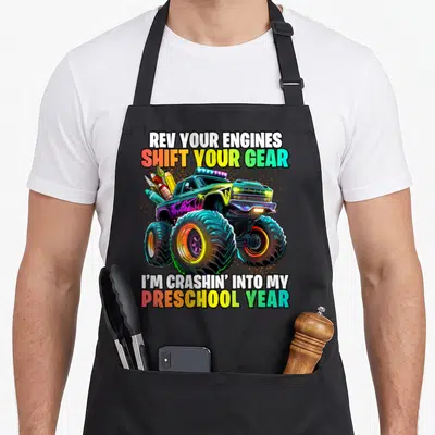 School preschool truck apron - Monster truck graphic with pencils and rainbow text Rev Your Engines Shift Your Gear — shown on a black cotton canvas apron against a white background