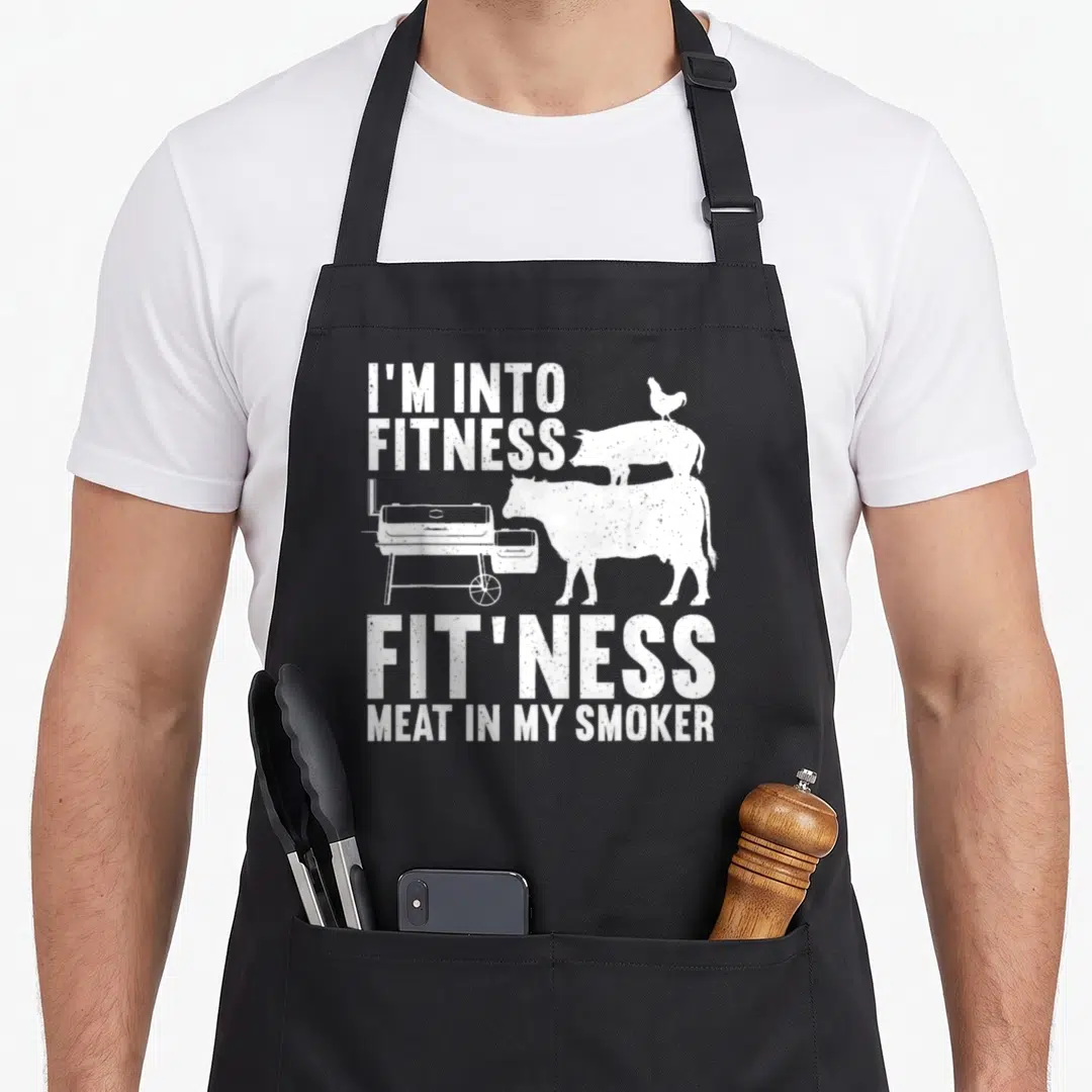 Meat smoking apron - White distressed text I'M INTO FITNESS over stacked cow pig chicken and smoker silhouette — shown on a black cotton canvas apron against a white background