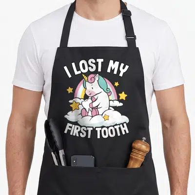 Lost unicorn apron - Cartoon unicorn holding a tooth on a cloud with rainbow and text I LOST MY FIRST TOOTH — shown on a black cotton canvas apron against a white background