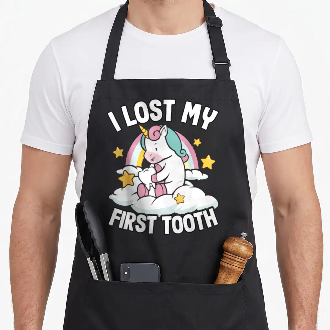 Lost unicorn apron - Cartoon unicorn holding a tooth on a cloud with rainbow and text I LOST MY FIRST TOOTH — shown on a black cotton canvas apron against a white background