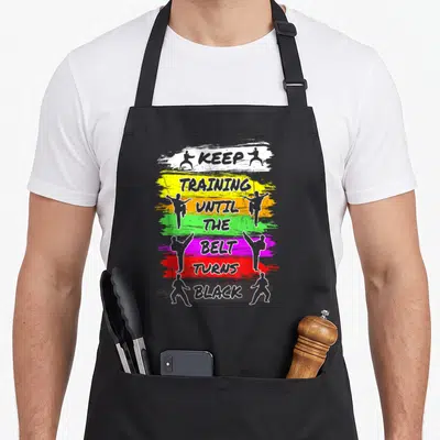 Training i turn grills on apron - Stacked text on colored brush strokes flanked by martial artist silhouettes in grey — shown on a black cotton canvas apron against a white background