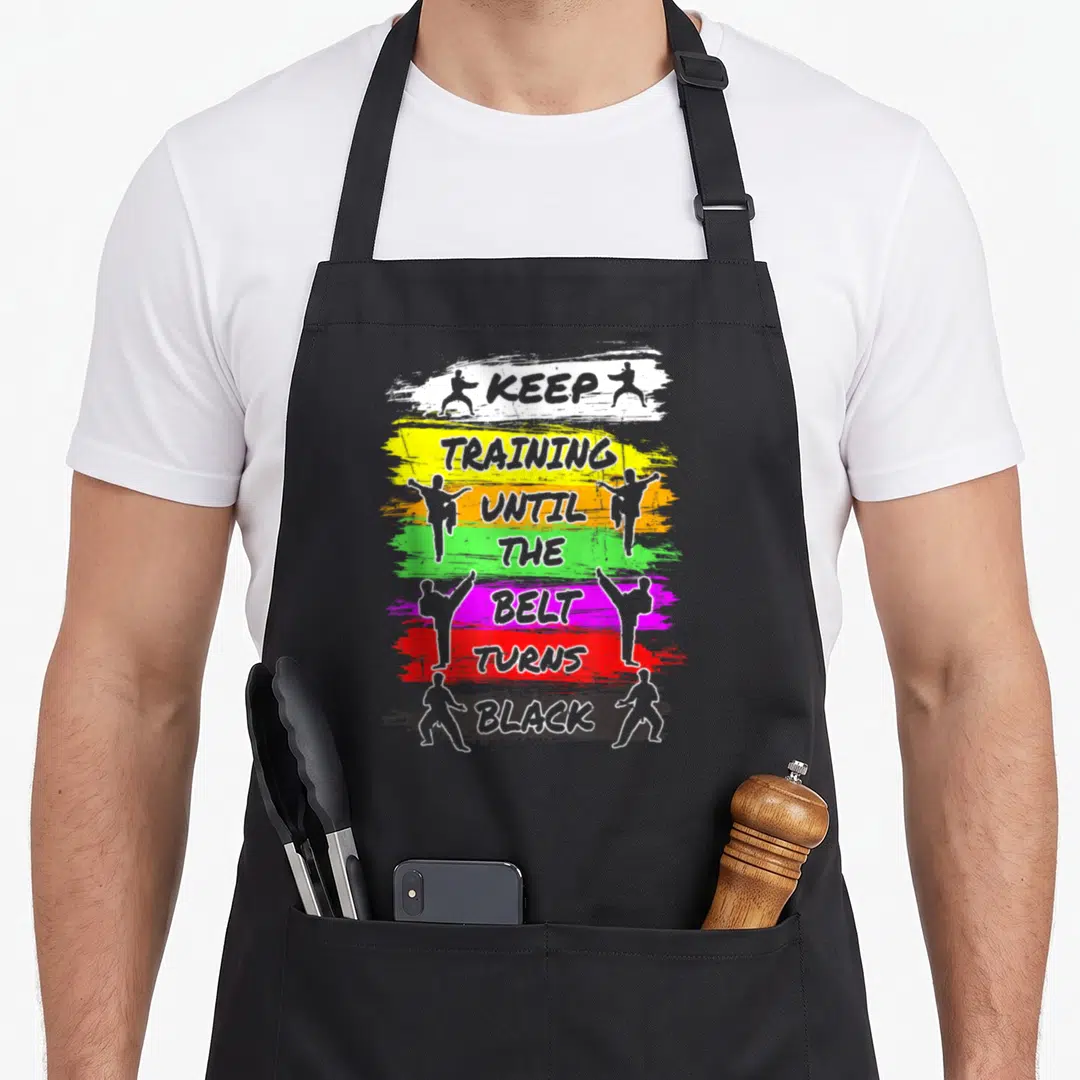 Training i turn grills on apron - Stacked text on colored brush strokes flanked by martial artist silhouettes in grey — shown on a black cotton canvas apron against a white background