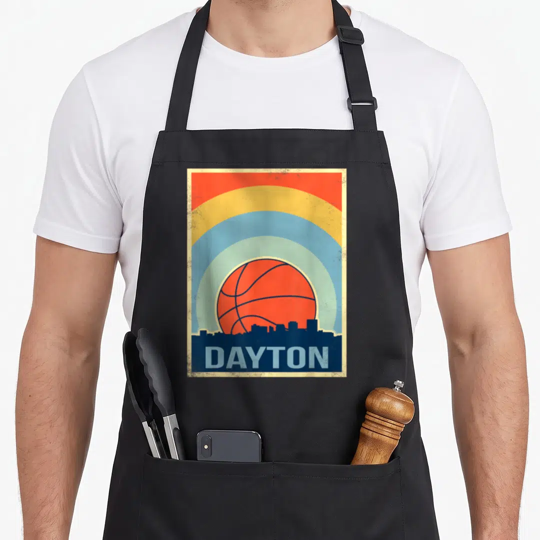 Funny basketball dayton apron - Orange basketball over dark blue Dayton skyline with red yellow blue arcs and DAYTON — shown on a black cotton canvas apron against a white background