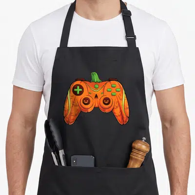 Gamer halloween kitchen aprons - Orange pumpkin game controller with green stem and jack-o-lantern face details — shown on a black cotton canvas apron against a white background