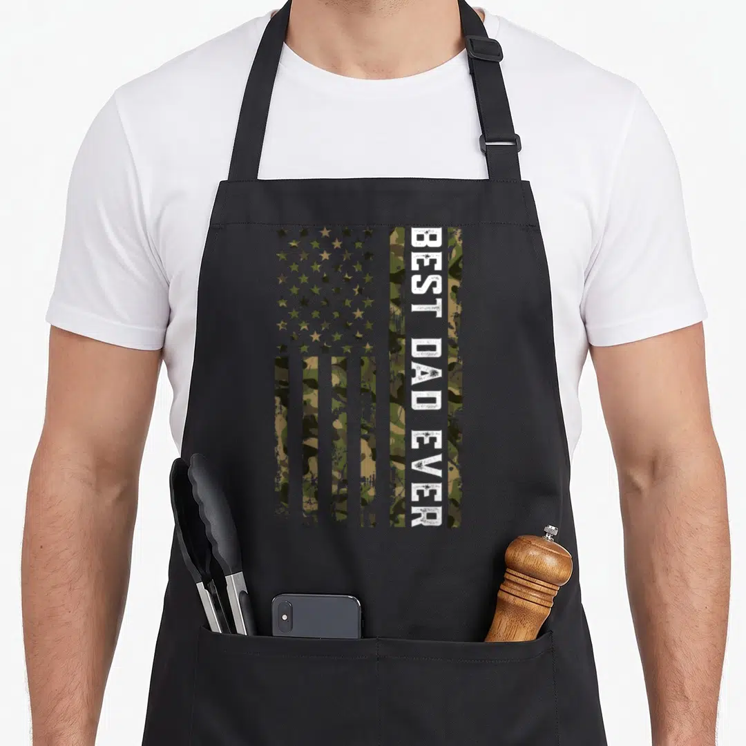 Patriotic camo apron - Camo USA flag with vertical white BEST DAD EVER text on distressed background — shown on a black cotton canvas apron against a white background