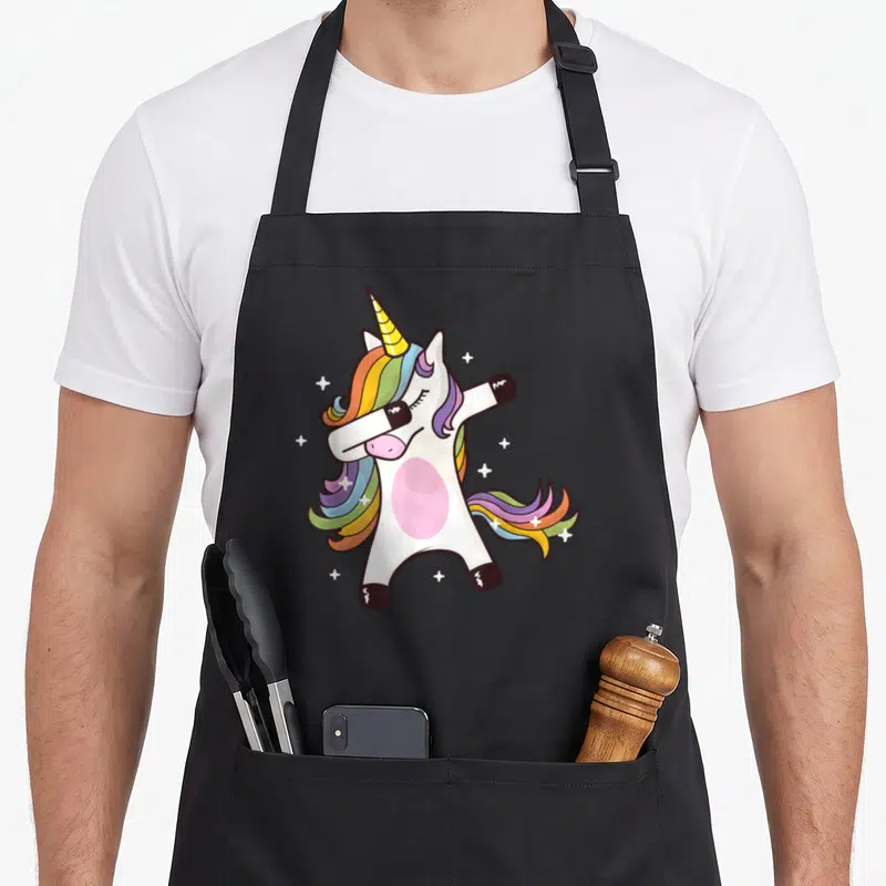 Pose unicorn apron - Cartoon white unicorn dabbing with rainbow mane and white stars — shown on a black cotton canvas apron against a white background