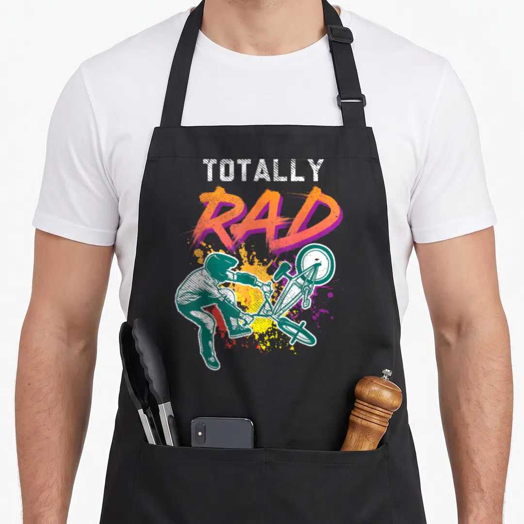 Bike 80s apron - Totally Rad text above green BMX rider and orange purple paint splatters — shown on a black cotton canvas apron against a white background
