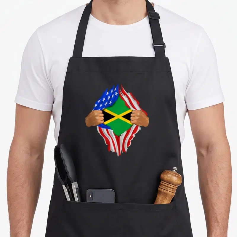 Jamaican blood apron - Two brown hands tearing USA flag to reveal Jamaican flag in blue red green yellow and brown — shown on a black cotton canvas apron against a white background