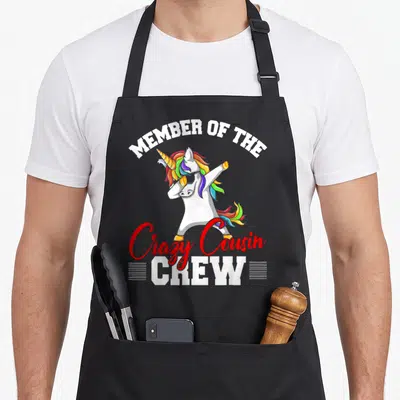 Member unicorn apron - Dabbing unicorn with rainbow mane wearing sunglasses under Member of the Crazy Cousin Crew text — shown on a black cotton canvas apron against a white background