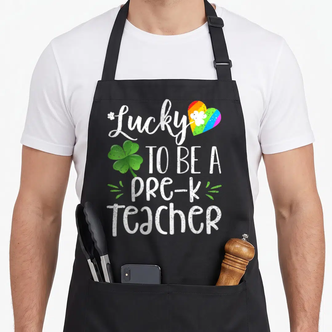 Pre st patrick day aprons - White text Lucky To Be A Pre-K Teacher with rainbow heart and green clover — shown on a black cotton canvas apron against a white background